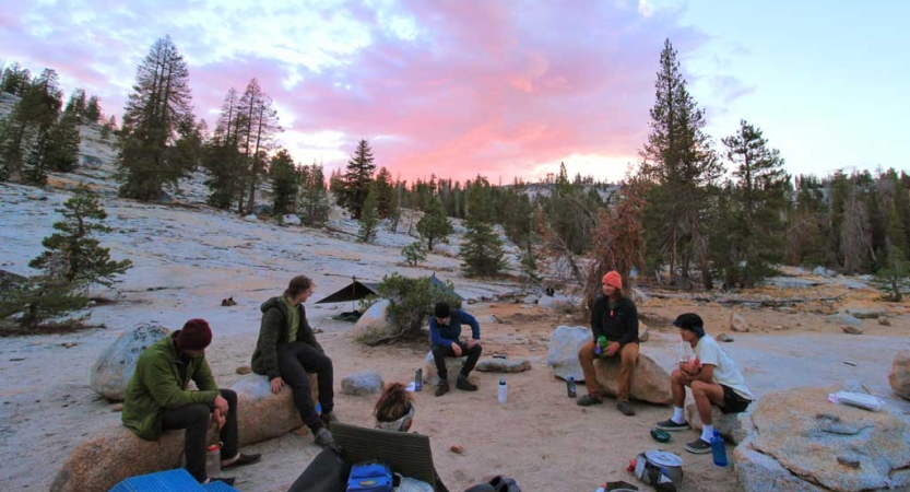 A group of people sit in a circle at camp. The sky appears in gentle shades of blue and pink as the sun either rises or sets. 